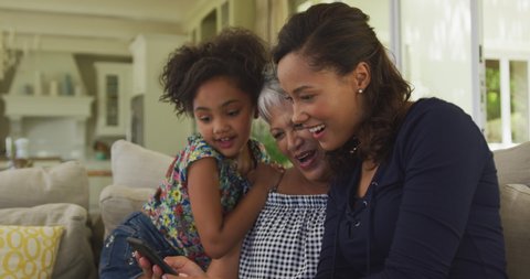 Senior African American woman, her adult daughter and her young granddaughter spending time at home together, sitting on a sofa in the living room, using a smartphone and smiling, in slow motion.