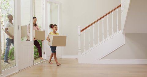 African American couple and their daughter moving into new house, walking through door and carrying cardboard boxes, smiling, in slow motion.
