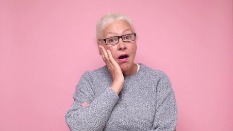 Sad old woman looking at camera showing distrustful look. Studio shot on pink wall.