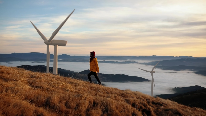 Epic shot of a woman hiking on the edge of the mountain against landscape with wind turbine power station on background. Concept of environmental engineering, renewable energy and love for nature. | Shutterstock HD Video #1061127232