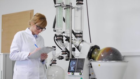 Scientist inspecting rotovapor in laboratory during CBD oil extraction. She is wearing rubber gloves and standing next to rotational vaporizer with green condenser.