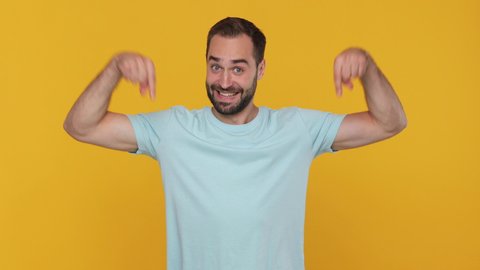 Fun expressive young man 20s years old in basic casual blue t-shirt look camera pointing fingers hands down on copy space workspace area isolated on yellow background studio. People lifestyle concept