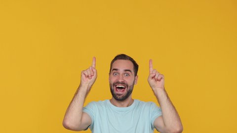 Shocked young man 20s years old in basic casual blue t-shirt look camera pointing fingers hands up overhead on copy space workspace area isolated on yellow background studio. People lifestyle concept