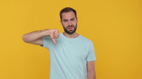 Bearded sad young man 20s in basic casual blue t-shirt isolated on yellow background studio. People emotions lifestyle concept. Looking camera showing pointing fingers down thumbs down gesture say no