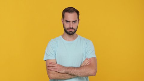 Bearded young man 20s in basic blue t-shirt isolated on yellow background studio. People sincere emotions lifestyle concept. Looking camera showing no finger gesture crossed folded hands disapproval