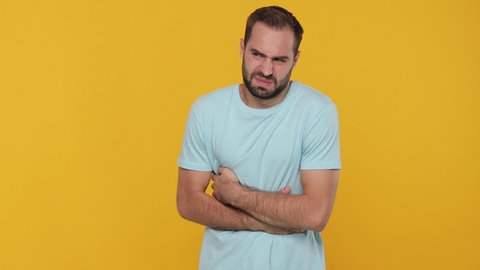 Bearded sad young man 20s in basic casual blue t-shirt looking camera feeling bad put his hands on stomach abdominal pain isolated on yellow background studio. People healthy ill sick disease concept