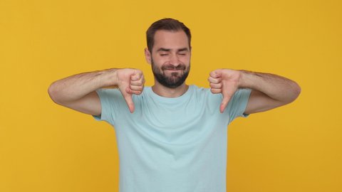 Bearded sad young guy 20s in basic casual blue t-shirt isolated on yellow background studio. People sincere emotions lifestyle concept. Looking camera showing pointing fingers down thumbs down gesture