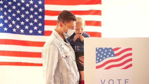 USA elections 2020 concept. Young man in a protective mask with a small child in his arms votes in a voting booth at a polling station