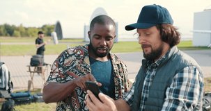 Portrait of multi-ethnic men standing outdoors and tapping and scrolling on smartphone. African American young man with happy Caucasian male browsing on cellphone and chatting. Backstage concept