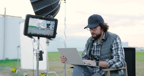 Handsome male film director in glasses and cap sitting in chair on backstage and typing on laptop outdoors. Caucasian man working and tapping on computer on set. Cinematography concept