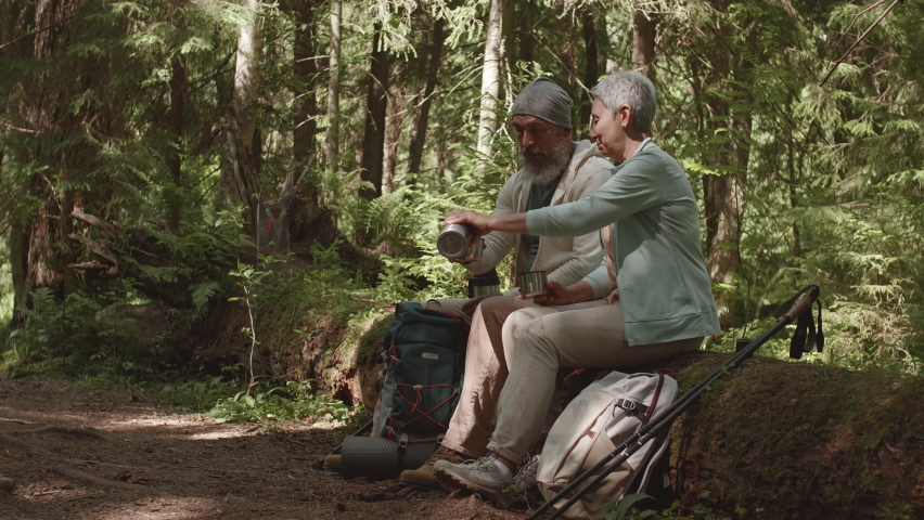 Panning of old multiethnic spouses are sitting on fallen tree in woods, resting and drinking tea from thermos bottle