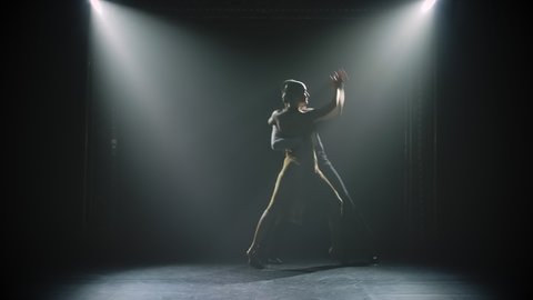 A young woman approaches her partner passionately and the couple dances Argentine tango. Dark silhouettes against the lights in the studio. Slow motion.