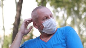 Video, an adult man in a blue T-shirt takes off and puts on a white protective mask. Outdoors in the park on a sunny day in summer.