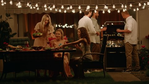 Cheerful friends enjoying barbeque party on backyard at evening. Handsome men preparing meat on grill. Attractive women talking at dining table near house. Young people spending time together outdoors