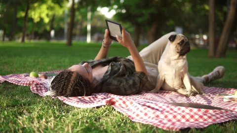 Woman laying on plaid on lawn in a park and reading e-book while little pug sitting next to her