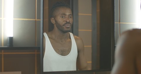 Reflection of young African American man looking in mirror and thinking. Thoughtful handsome guy with tattoo on hand leaving bathroom in the morning.