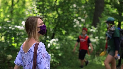 Beautiful female wearing covid-19 protective face mask to fight against coronavirus with a side bag walking in park with children passing by