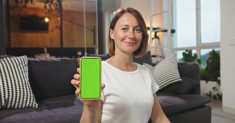 Middle plan of happy woman sitting on floor holding phone with chromakey. Cute female showing phone with a green screen. Concept modern technologies, chroma key, smartphone, cell phone, lifestyle.