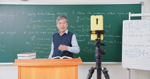 Asian senior male calculus professor is ponting on board while having blackboard lecture online through smartphone in the classroom at college