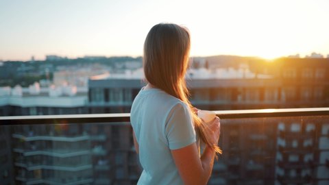 Woman with a cup of coffee or tea goes to the balcony to admire the sunset