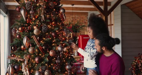 Loving african father and little daughter talking standing near ornate twinkle lights Christmas tree. New Year preparation and celebration, winter holiday make festive spirit at modern home concept