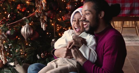 African family playing in modern living room decorated with twinkle lights and Christmas tree, 30s father hugging tickling little son people laughing enjoy funny time together. Happy holidays concept