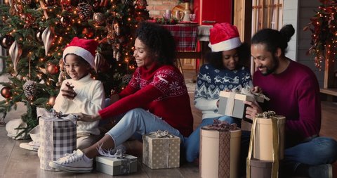 Loving parents helping to African kids in Santa hats open gifts, full family sit together near decorated glowing garland Christmas tree enjoy moment of surprises unboxing. New Year celebration concept