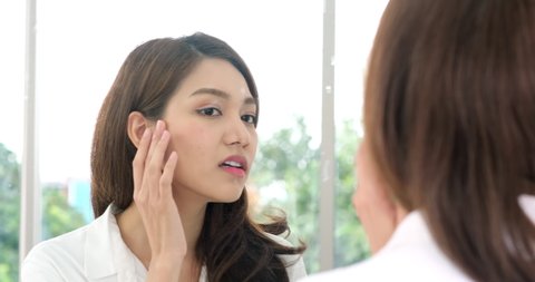 Portrait of worried young Asian woman looking in mirror, examining acne and wrinkles on her face. Women never applied facial cream Until face has wrinkles and acne.