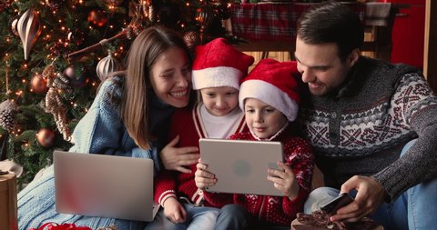 Family with small kids gathered at home near glowing Christmas tree spend holidays time using diverse electronic devices. Overuse, parental control, New Year celebration, gifts purchase online concept