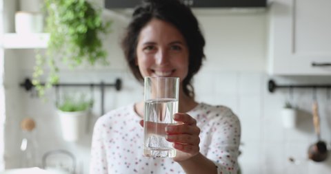 Smiling young woman standing in home kitchen stretch out hand to camera glass of natural still water, offers you clean aqua, close up focus view. Healthy life habits, healthcare and lifestyle concept