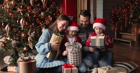 Happy family with little son and daughter in Santa hats on x-mas morning open gifts boxes sit near glowing Christmas tree in cozy ornate living room at modern luxury home. New Year celebration concept