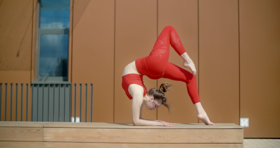 Gymnastic woman in red does acrobatic exercices on the bench in the yard of an apartment building, fitness in the urban environment, callisthenics outdoors, physical workout, 4k 120p Prores