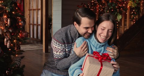 At decorated hall modern house young couple sit near xmas tree celebrating New Year, husband make Christmas surprise for wife closing her eyes with hands giving gift box. Happy winter holidays concept