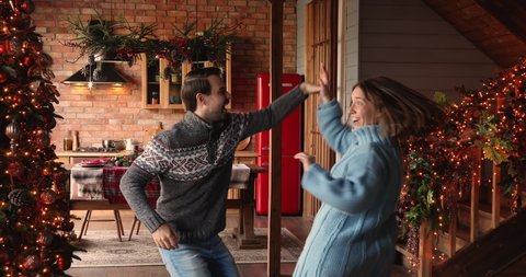 Happy young active couple in winter sweaters enjoy Christmas Eve and 2021 New Year winter holidays dancing indoor at ornate twinkle lights xmas tree home hall. Fun and hobby, x-mas celebration concept