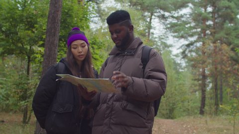 Irritated lovely interracial hikers couple with backpacks got lost in forest, looking at travel map and arguing about right route trek, pointing in different directions during hiking in autumn nature.