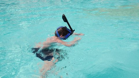 An unrecognizable boy floats on the blue surface of the water in an underwater mask with a snorkel in slow motion in a resort pool.