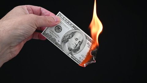 Burning a US $100 bill. A hand holds the banknote while lights it on fire using a lighter. Black background, slow motion.
