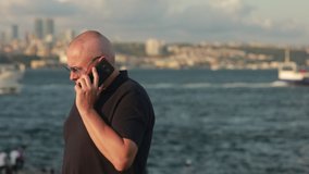 Video, an emotional adult man in a polo shirt and sunglasses is talking on a smartphone. On a sunny day in summer, against the backdrop of the blue sea and sailing ships.
