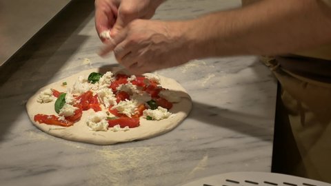 Preparing Pizza Margherita on a marble countertop. Pizzaiolo puts fresh tomatoes, mozzarella and basil leaves over a raw pizza dough. 