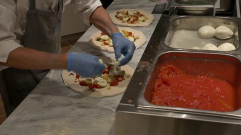 Preparing Pizza Margherita on a marble countertop. Pizzaiolo puts fresh tomatoes, mozzarella and other ingredients over a raw pizza dough. 