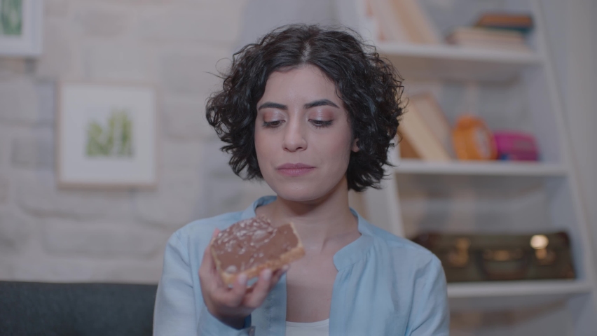 Young woman is sitting on an armchair at home and eating chocolate bread. | Shutterstock HD Video #1061178325