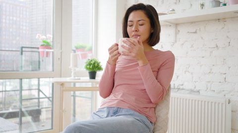 Asian mix race woman enjoying cup of coffee or tea drinking warm beverage at home. Young girl relaxing with morning drink in hands looking through window enjoying and dreaming