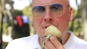 Video, an emotional adult man in a white shirt and sunglasses eats fresh fig fruit in a public park. On a sunny day in summer.