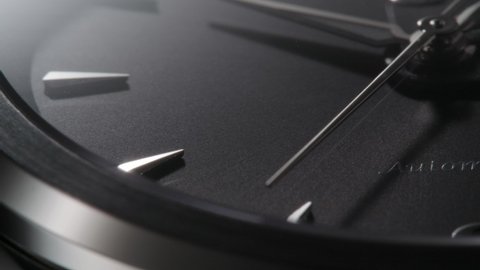 Extreme closeup of metallic second arrow passing by on black clock face. Lengthwise view of luxury swiss watch 
