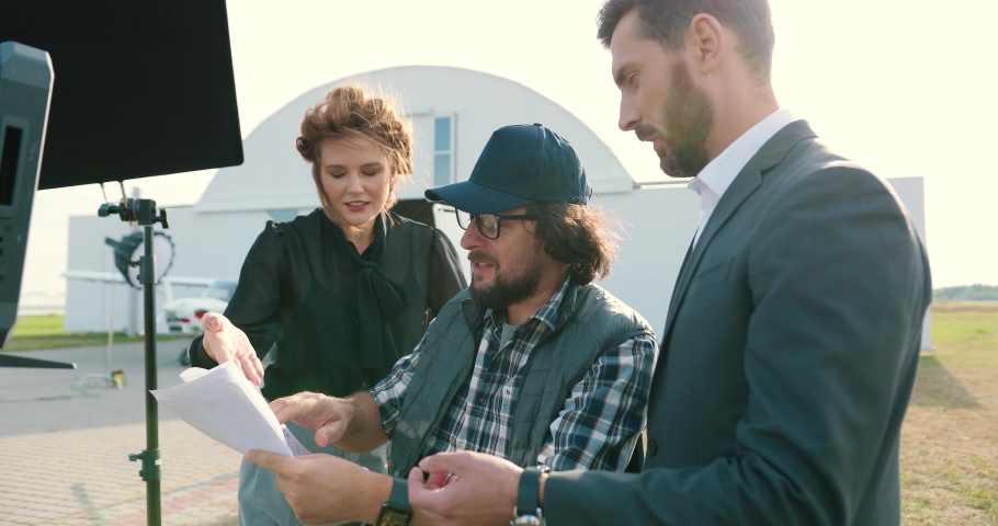 Portrait of film crew members speaking outdoors on set. Handsome Caucasian male and beautiful female chatting with movie director on backstage. Filmmaker discussing script with actors. Filming concept Royalty-Free Stock Footage #1061181280