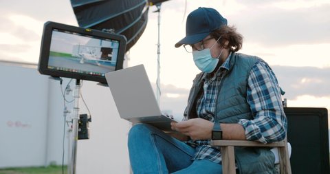 Portrait of Caucasian male filmmaker in mask and glasses sitting into director chair and looking into screen on set while shooting video outdoors. Man with laptop on backstage. Cinematography concept