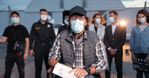 Portrait of man in mask and cap sitting in director chair on set outdoors in spotlight. Mixed-race film crew on background. Males and females standing in masks after shooting. Cinematography concept