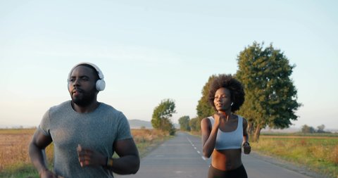 Cheerful African American young sporty man and woman joggers in headphones running on road early in the morning in countryside. Couple of sportsman and sportswoman jogging and listening to music.
