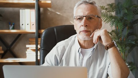 Mature businessman in glasses is thinking and typing on laptop while sitting at desk working at home. Palm tree, shelving with books and ring binders on background. Remote job, freelance. Close up