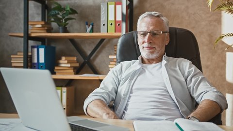 Mature man in glasses is leaning back in armchair, having rest being satisfied about done work at home office. Sitting at desk with laptop and notepad. Remote job, business. Close up, slow motion